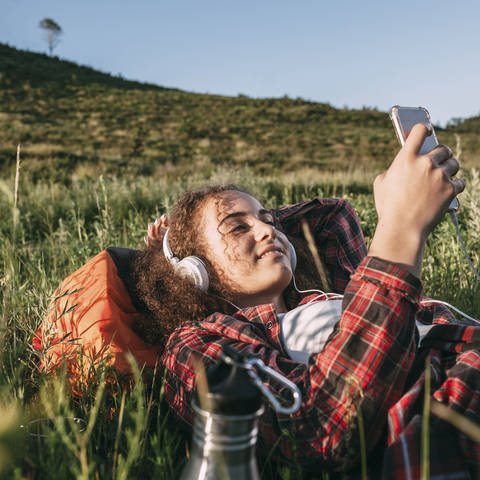 Eine junge Frau liegt im Gras und hört Podcast (Foto: IMAGO, Imago/Vasily Pindyurin)