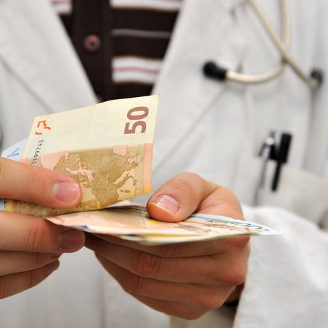 Ein Mann im Arztkittel zählt Geldscheine (Foto: dpa Bildfunk, Picture Alliance)