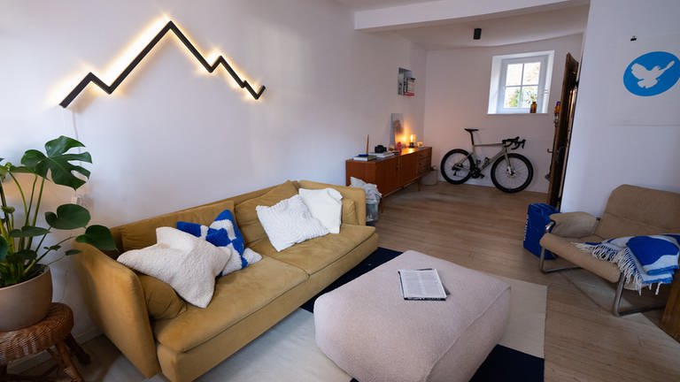Ein gemütliches Wohnzimmer mit einer senfgelben Ikea-Couch, Kissen und einer modernen, auffälligen Wand-Lampe in Form eines Bergmassivs (Foto: SWR)