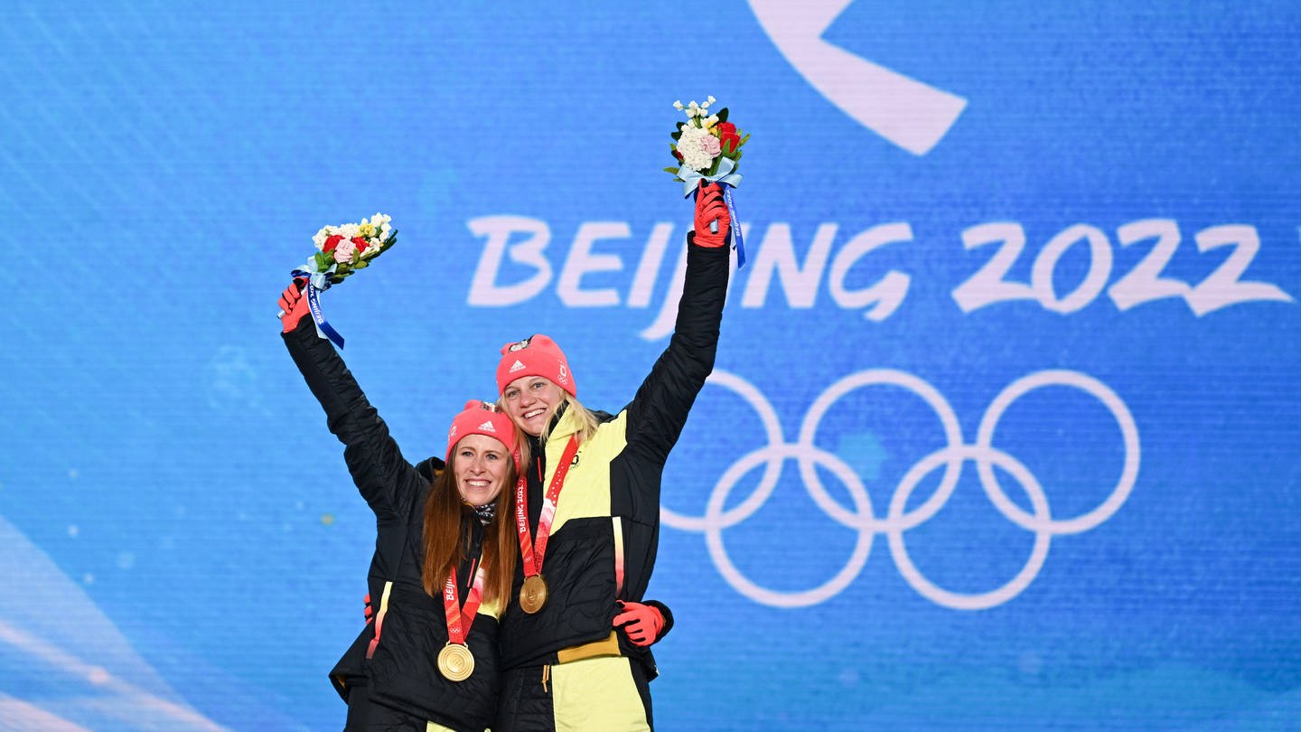 Katharina Hennig (l.) und Victoria Carl (r.) stehen bei der Siegerehrung vor einer blauen Wand, auf der Beijng 2022 steht. Um den Hals tragen sie Goldmedaillen und sie recken Blumensträuße in die Luft. (Foto: IMAGO, Xinhua)