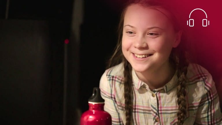 Standbild aus dem Dokumentarfilm Ich bin Greta: Greta sitzt lächelnd vor einem Notebook, rotes Overlay mit Icon Kopfhörer (Foto: SWR, WDR, B-Reel Features Film)