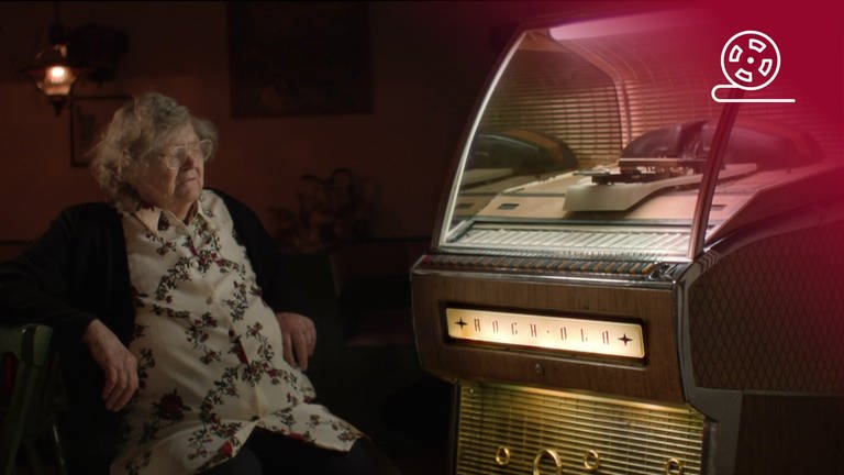 Standbild aus dem Dokumentarfilm 80.000 Schnitzel: Berta Zenefels sitzt vor einer Jukebox, rotes Overlay mit Icon Filmrolle (Foto: SWR, Zum goldenen Lamm, ZDF, Stefanie Reinhard)