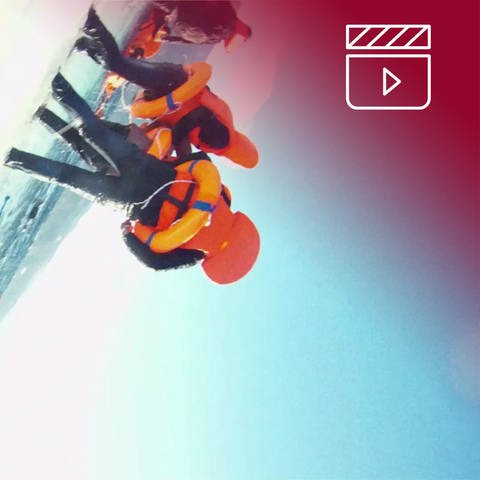 Standbild aus dem Dokumentarfilm Das Purpurmeer: Menschen mit Rettungswesten teils am Strand stehend, teils im Mittelmeer, treibend rotes Overlay mit Icon Filmklappe (Foto: SWR, pong film, Amel Alzakout)