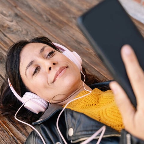 SWR1 zum Hören, wann und wo Sie wollen (Foto: Frau mit Kopfhörern und Smartphone liegt auf dem Holzboden) (Foto: IMAGO, IMAGO / Addictive Stock)