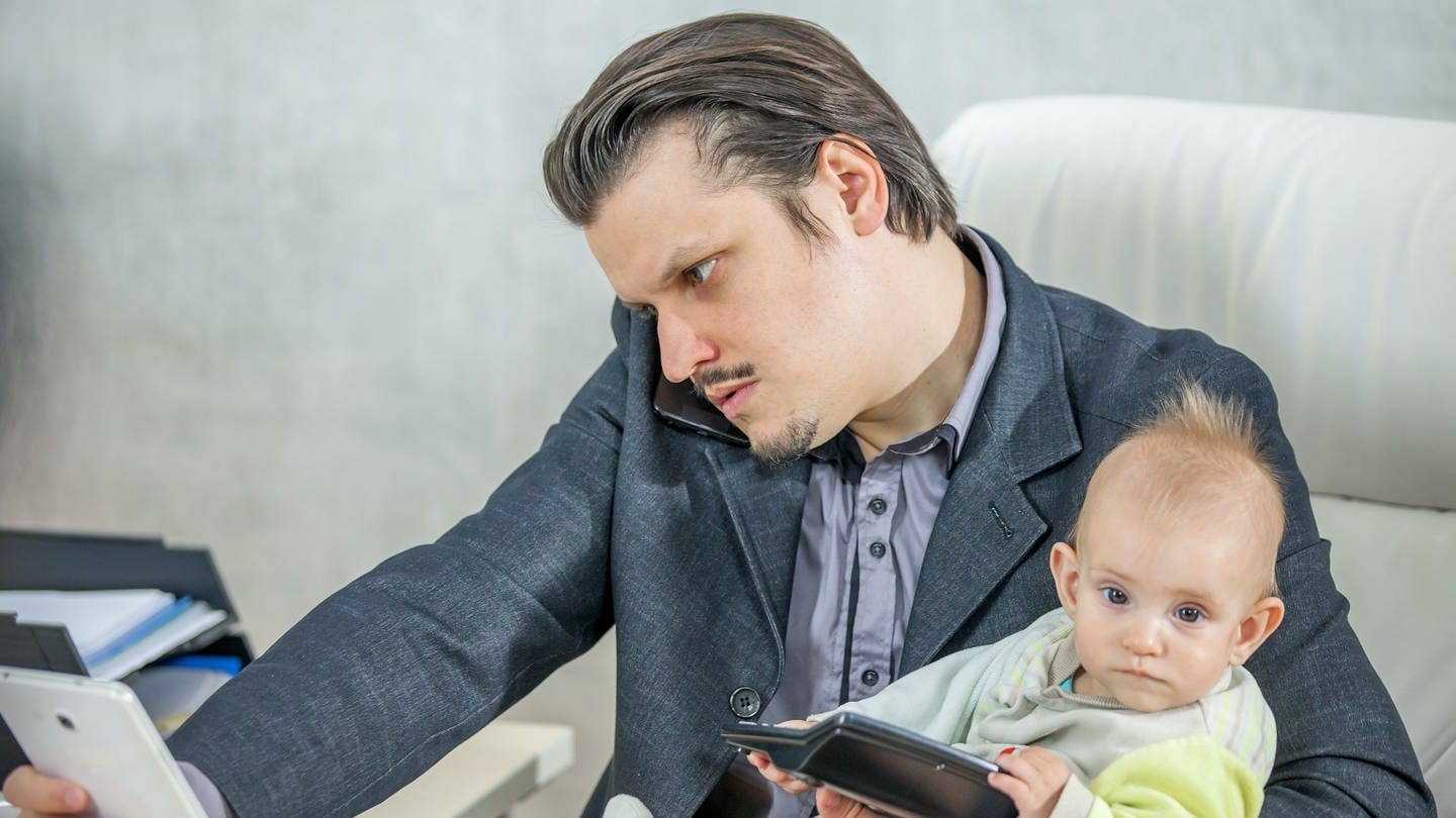 Ein junger Geschäftsmann arbeitet in seinem Büro und hält ein Baby (Foto: IMAGO, IMAGO / Wirestock)
