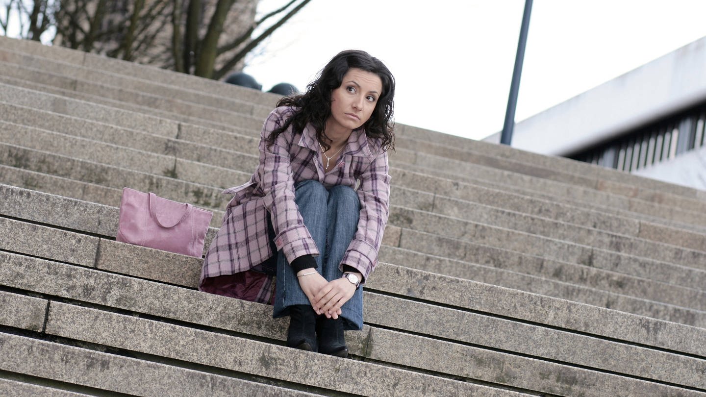 Junge Frau sitzt gelangweilt auf einer Treppe. (Foto: IMAGO, IMAGO / blickwinkel)