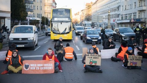 Aktivisten der Klimaschutzgruppe Letzte Generation sitzen bei einer Straßenblockade mit Schildern mit aufgemalten Traktoren auf der Straße. Aktivisten nehmen damit Bezug auf die Aktionswoche der Landwirte. (Foto: picture-alliance / Reportdienste, picture alliance/dpa | Sebastian Gollnow)