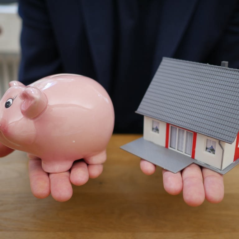 Eine Person streckt seine beiden Hände nach vorne, darauf links ein Sparschwein, rechts das Modell eines Einfamilienhauses. (Foto: unsplash.com/Tierra Mallorca)