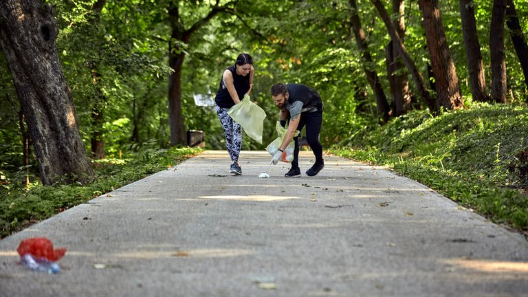 Ein Mann und eine Frau sammeln Müll im Wald. Ploggin ist eine Wortzusammensetzung aus dem schwedischen Wort "plocka" und joggen. Es bedeutet: beim Joggen Müll aufsammeln und damit etwas für den Umweltschutz tun. Sport und Fitness Training plus Naturschutz.
