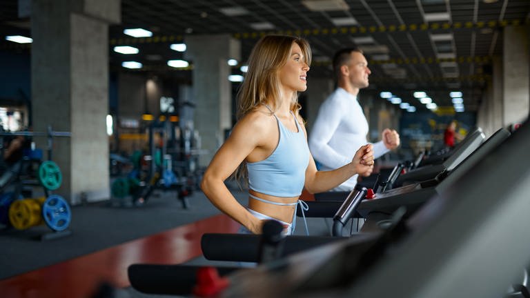 Eine Frau und ein Mann laufen auf Laufbändern im Fitnessstudio. 10.000 Schritte, Muskelkater ist gut, Joggen zum Abnehmen und gezielte Fettverbrennung: Sportwissenschaftlerin Dr. Mona Kellner klärt Mythen zu Sport und Ernährung auf.