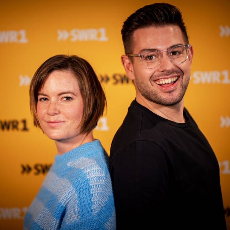 Die SWR1 Moderatoren Cora Klausnitzer und Matthias Sziedat stehen mit dem Rücken aneinander und lächeln in die Kamera. (Foto: SWR)