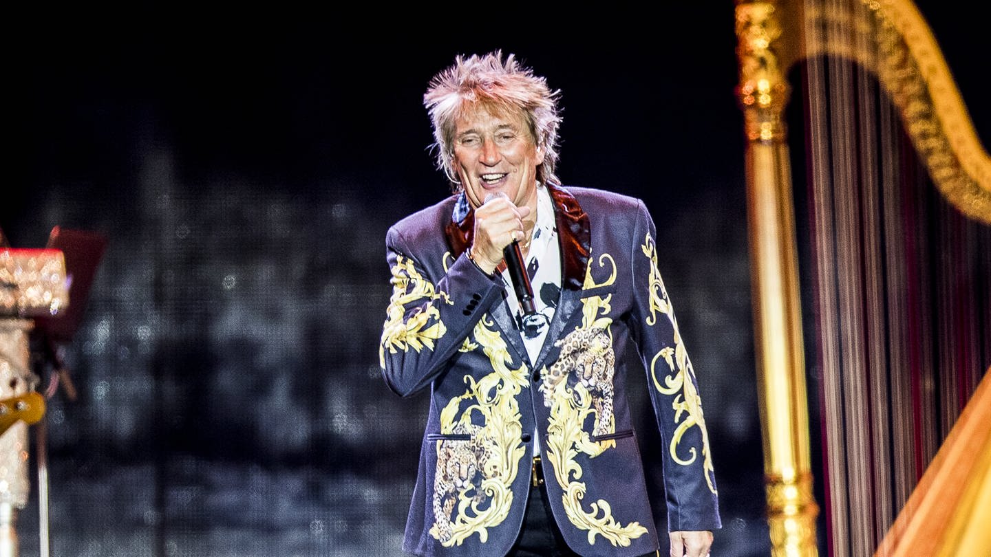 Der britische Sänger Rod Stewart trägt einen dunkelblauen Anzug mit goldenem Muster. Er singt in ein schwarzes, kabelloses Mikrofon. (Foto: dpa Bildfunk, Picture Alliance)