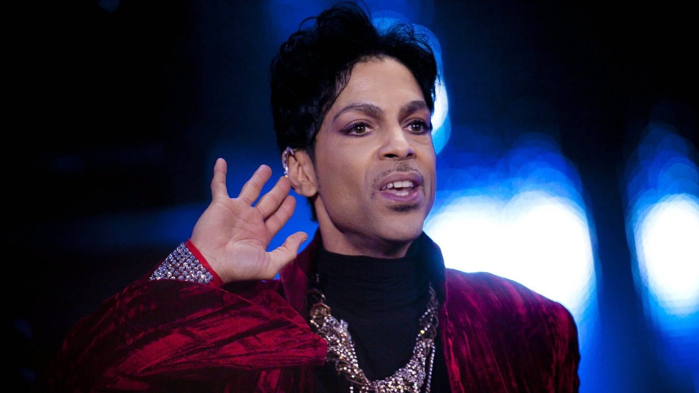 Der Sänger Prince steht auf einer Bühne. Er trägt ein rotes Samtoberteil mit glitzernden Ketten. (Foto: dpa Bildfunk, Picture Alliance)