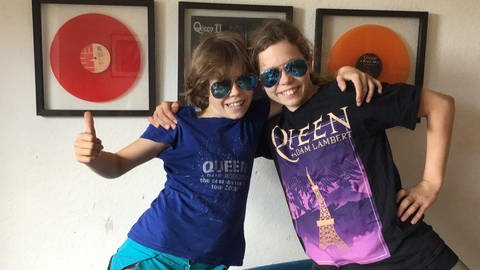 Hörernachricht mit einem Selfie von zwei Kindern mit Queen-Merch Tshirts an. Beide Kinder haben Sonnebrillen auf. Im Hintergrund hängen Queen Schallplatten