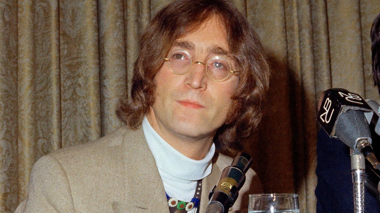 John Lennon in einem grau-beigen Anzug vor einer grau-beigen Gardine