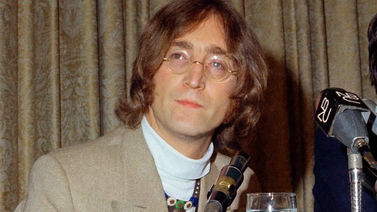 John Lennon in einem grau-beigen Anzug vor einer grau-beigen Gardine