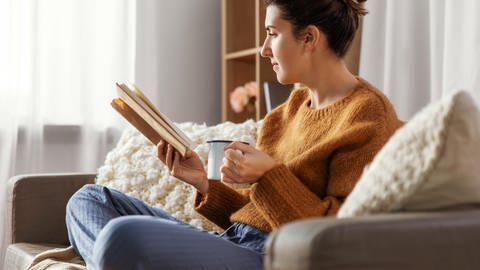 Eine Frau sitzt im Schlafanzug auf einer Couch und liest ein Buch. In der Hand hält sie eine Tasse Kaffee. (Foto: picture-alliance / Reportdienste, Picture Alliance)