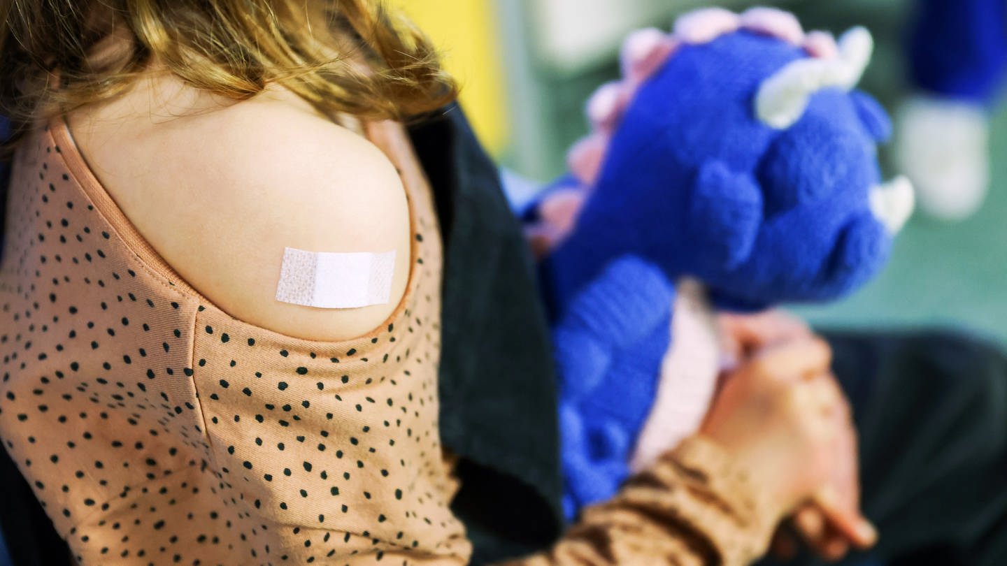 Ein Kind hält einen blauen Dino als Kuscheltier im Arm. An ihrem Arm hat sie ein Impfpflaster.