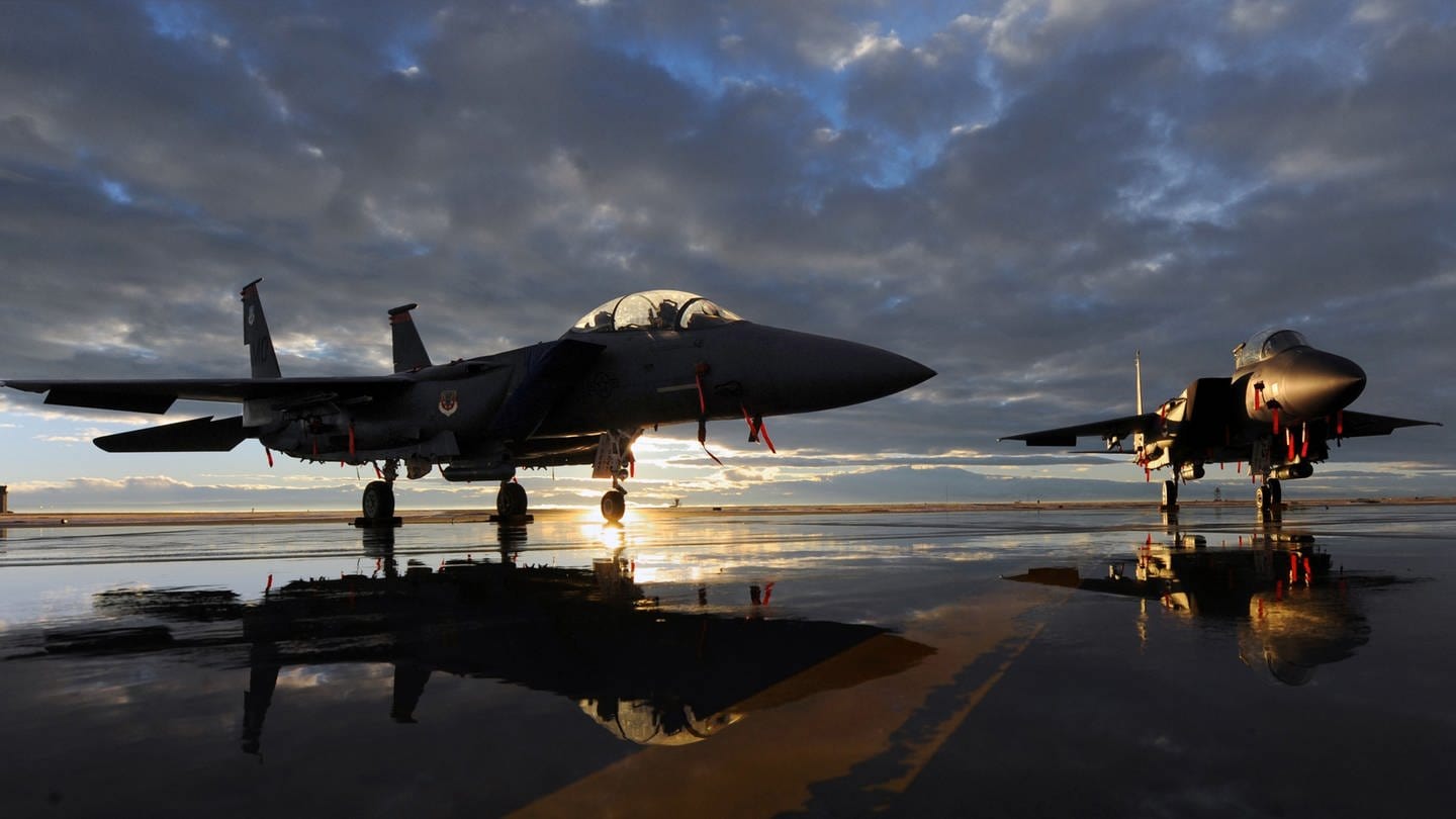 Ein Kampfjet, ein F-15 Strike Eagle fighter, steht zwischen Pfützen auf einem Rollfeld im Sonnenuntergang, im Hintergrund türmen sich dunkle Wolken auf. (Foto: picture-alliance / Reportdienste, picture alliance / Everett Collection)