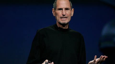 Steve Jobs steht auf einer dunklen Bühne und trägt eine schwarze Jacke. Er streckt beide Arme von sich und präsentiert etwas. (Foto: picture-alliance / Reportdienste, picture alliance / dpa | Monica M. Davey)