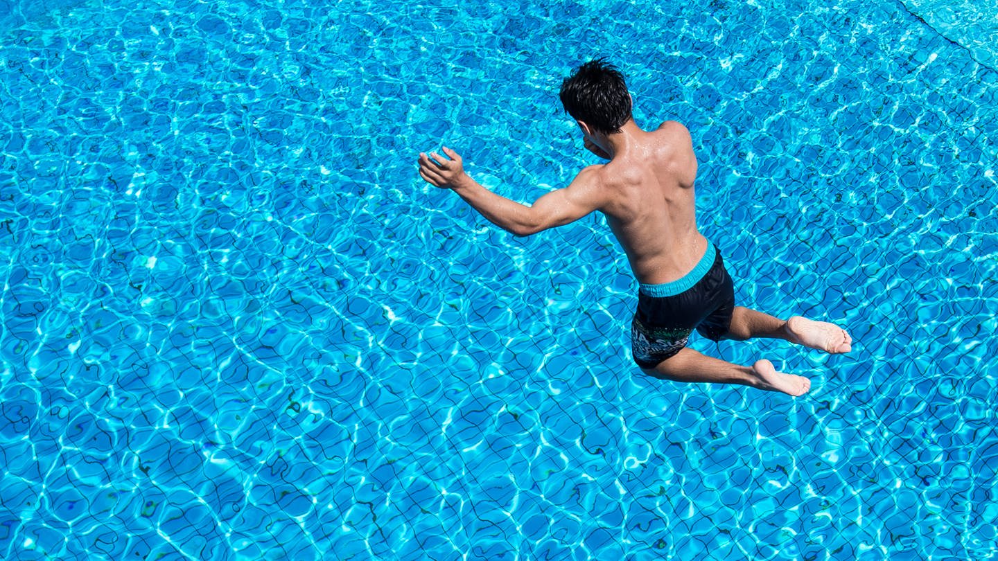 Ein Junge springt in einem Freibad von einem Sprungturm ins Wasser.