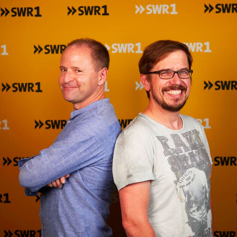 Die SWR1 Moderatoren Patrick Neelmeier und Ingo Lege stehen mit dem Rücken aneinandergelehnt und lächeln in die Kamera. (Foto: SWR)