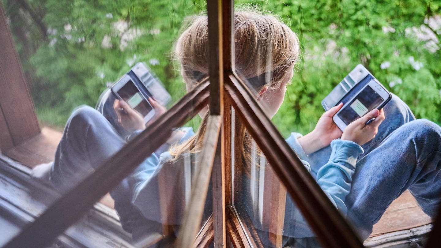 Ein Kind sitzt am Fenster und schaut auf sein Handy (Foto: picture-alliance / Reportdienste, picture alliance/dpa | Annette Riedl)