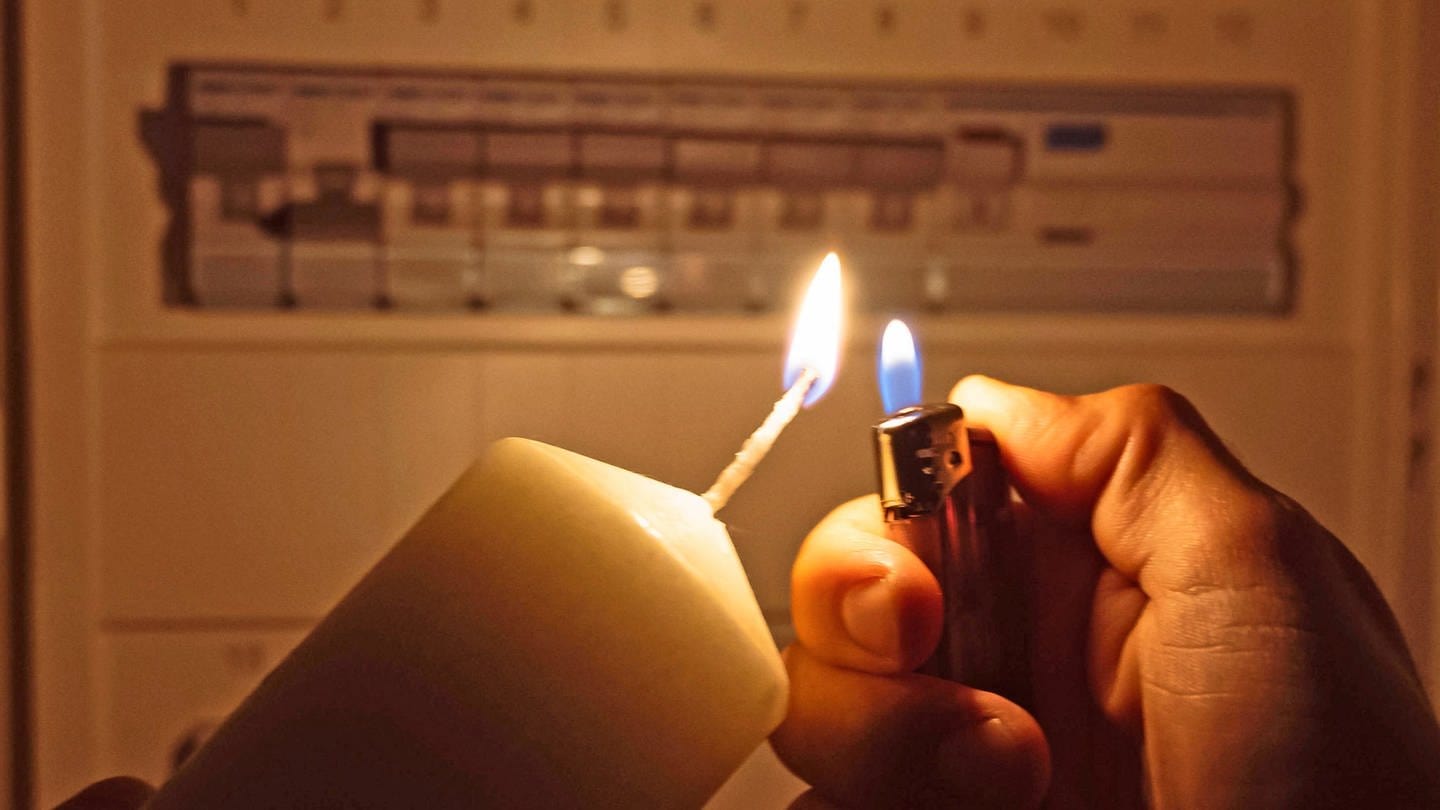 Stromausfall: ein Mann zündet mit einem Feuerzeug eine Kerze an, dahinter sieht man einen Sicherungskasten - die Sicherungen sind offensichtlich alle in 