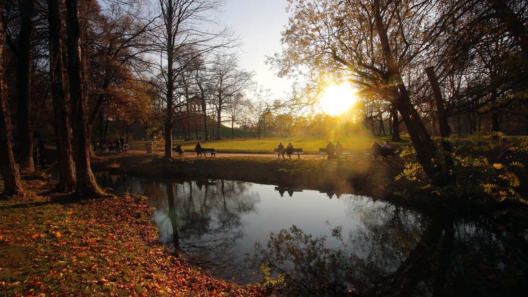 Ein Stadtpark mit einem kleinen See, indem sich die Sonnenstahlen spiegeln. Menschen sitzen auf Bänken und genießen die Natur. (Foto: IMAGO, Ralph Peters)