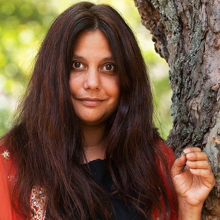 Mithu Sanyal schaut selbstbewusst in die Kamera. Sie trägt ein schwarzes Kleid und einen roten Überwurf mit goldenen Blüten  (Foto: Guido Schiefer/ Hanser Verlag)