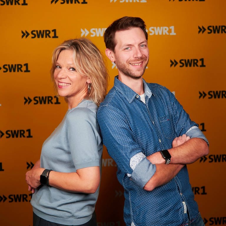 Die SWR1 Moderatoren Stefanie Anhalt und Corvin Tondera-Klein stehen mit dem Rücken aneinandergelehnt und lächeln in die Kamera. (Foto: SWR)
