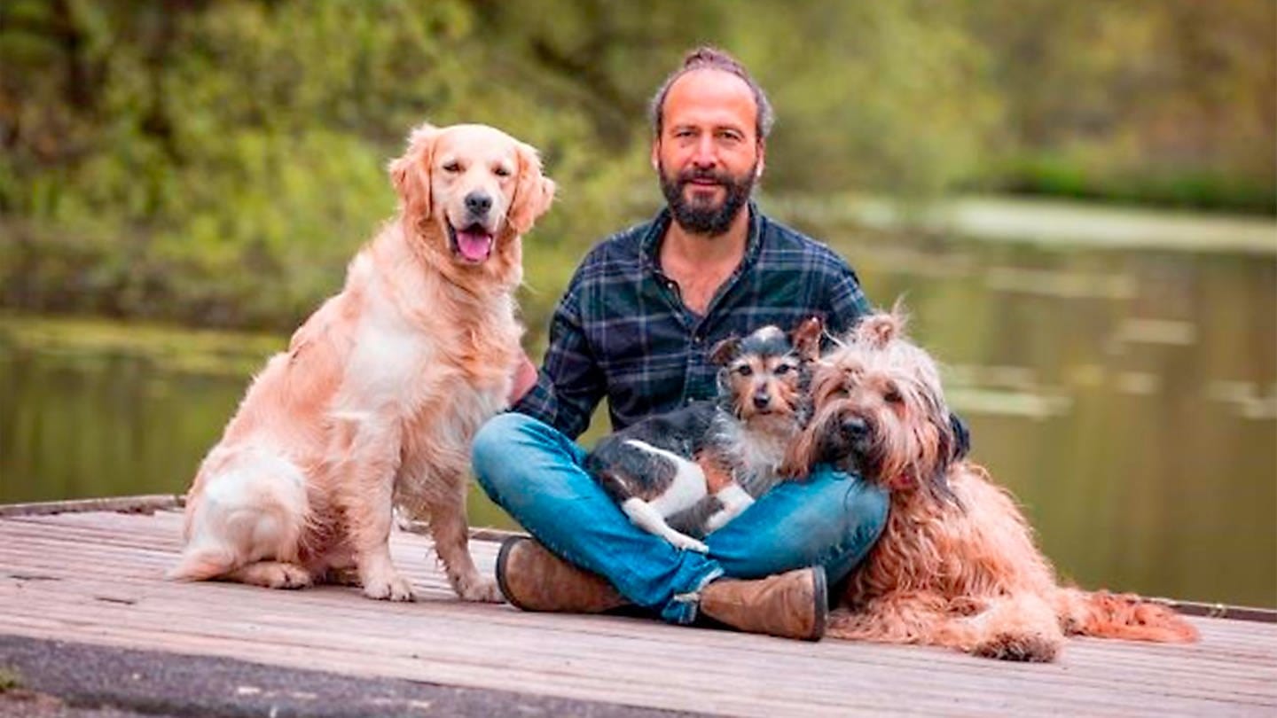 Ein Mann sitzt mit drei Hunden auf einem Holzsteg an einem Gewässer. Trainer, Dozent und Jurist Sami El Ayachi hat eine spannende andere Form des Hundetrainings entwickelt, die er uns im Podcast 