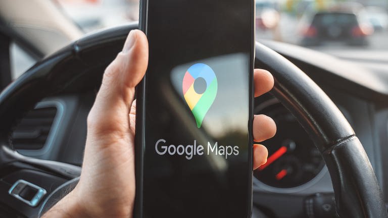 Eine Hand hält ein Smartphone mit dem Logo von "Google Maps" auf dem Bildschirm, im Hintergrund Lenkrad und Tacho eines Autos