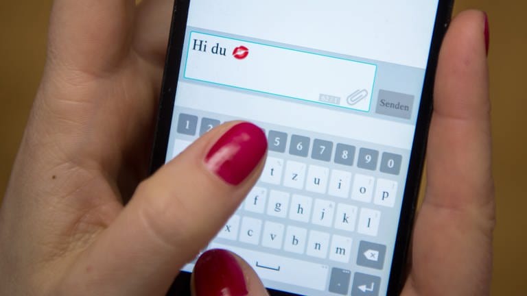 Eine Frau tippt auf einem Handy die Worte "Hi du" mit einem Kussmund-Emoji in einen Chat. Love oder Romance Scamming ist Betrug auf Online Dating Plattformen. So schützt Ihr Euch vor Liebesbetrug im Internet: Love Scam erkennen - das raten Polizei und Psychologen