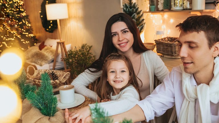 Eine junge Familie sitzt gemütlich und zufriedend lächelnd an einem Tisch inmitten von Weihnachtsdekoration (Foto: picture-alliance / Reportdienste, picture alliance / Zoonar | Dasha Petrenko)