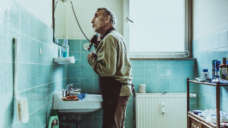 Fotowerk von Andy Reiner: Ein Mann mit einer Schürze um steht im Badezimmer vor dem Spiegel und rasiert sich mit einem Elektrorasierer. (Foto: Andreas Reiner / Sichtlichmensch Fotografie)