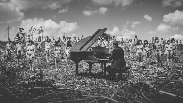 Fotowerk von Andy Reiner: Ein Pianist spielt im Freien an einem Konzertflügel, hinter ihm stehen viele unbekleidete Frauen und Männer. (Foto: Andreas Reiner / Sichtlichmensch Fotografie)