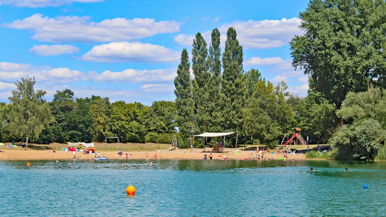 Ein See mit Booten und Surfern, Strand und Bäumen in der Sonne: Das ist der Hardtsee in Ubstadt-Weiher bei Bruchsal.
