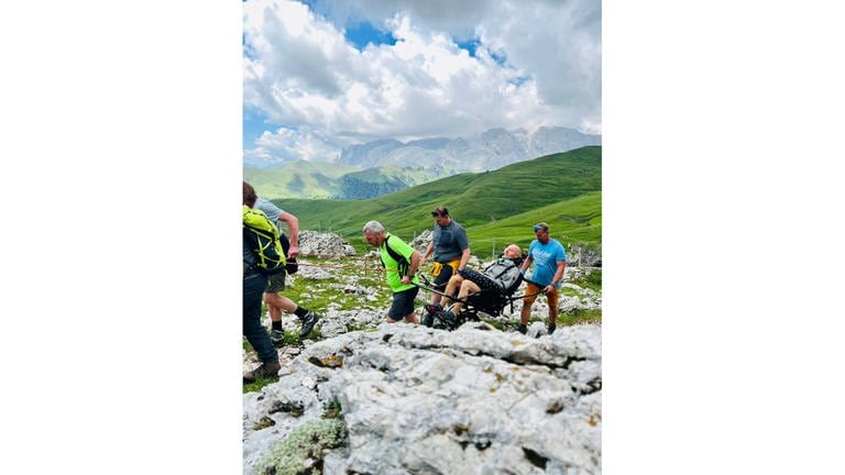Mehrere Männer ziehen und tragen einen Mann im Rollstuhl durch die Dolomiten. Stefan Kappler hat ALS und kann deshalb nicht mehr in den Bergen wandern. Seine Freunde machen mit ihm die ersehnte Tour durch die Dolomiten. Wie er es schafft, trotz der Amyotrophe Lateralsklerose (ALS) seine Lebensfreude zu behalten, erzählt er in der Audioreihe  Podcast SWR1 Neuanfang.