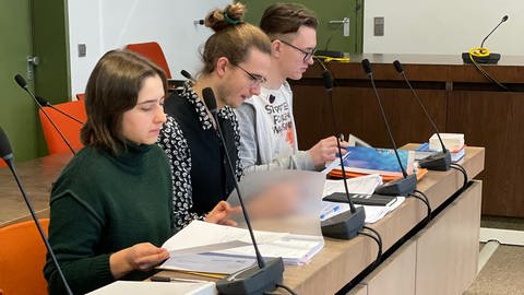 Maria, Moritz und Malte - die drei Aktivisten der "Letzten Generation" auf der Anklagebank. Sie sitzen an einem Tisch mit Mikrofonen und blättern in Aktenordnern. Sie stehen wegen einer Aktion vor Gericht, bei der sie für die Letzte Generation teilgenommen haben.