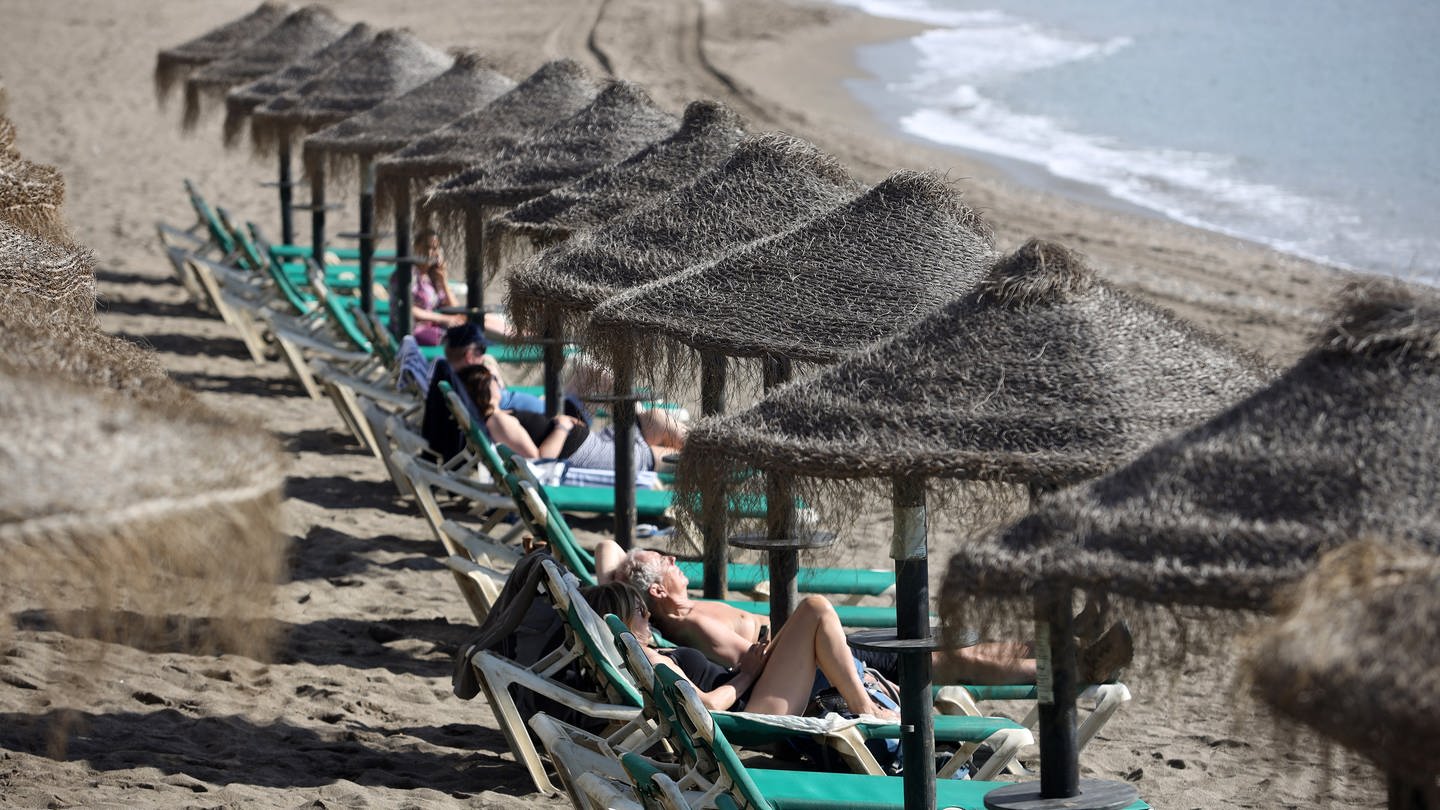 Menschen liegen am Strand von Spanien auf Strandliegen mit Sonnenschirmen aus Stroh (Foto: picture-alliance / Reportdienste, picture alliance/dpa/EUROPA PRESS | Álex Zea)