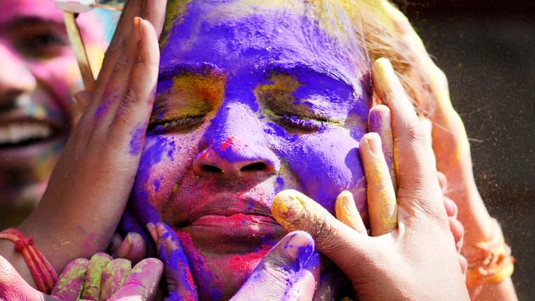 28. März: Das Leuchten des Frühlings! Eine junge Frau schließt die Augen und feiert auf dem Holi Festival, dem Fest der Farben. Ihr Gesicht - umzingelt von Händen - ist bedeckt mit Neonpulver in lilanen, pinken und gelben Tönen.  (Foto: picture-alliance / Reportdienste, picture alliance/dpa/AP|Rajesh Kumar Singh)