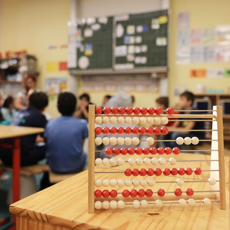 Ein Rechenschieber steht auf einem Tisch im Klassenzimmer in einer Grundschule. Im Hintergrund sind die Tafel und Kinder zu erkennen. (Foto: dpa Bildfunk, picture alliance/dpa | Oliver Berg)