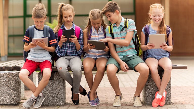 Fünf Schulkinder sitzen auf einer Bank und schauen auf ihre Tablets