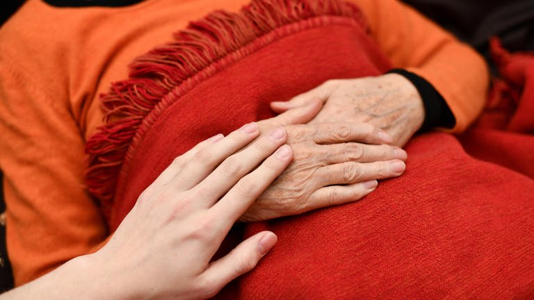 Frau liegt mit gefalteten Händen im Bett, eine andere Hand streicht die ihre