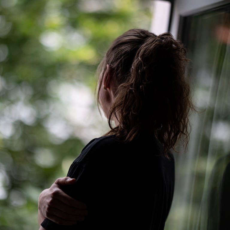 Eine Frau steht in einer Wohnung und schaut durch ein Fenster nach draußen.