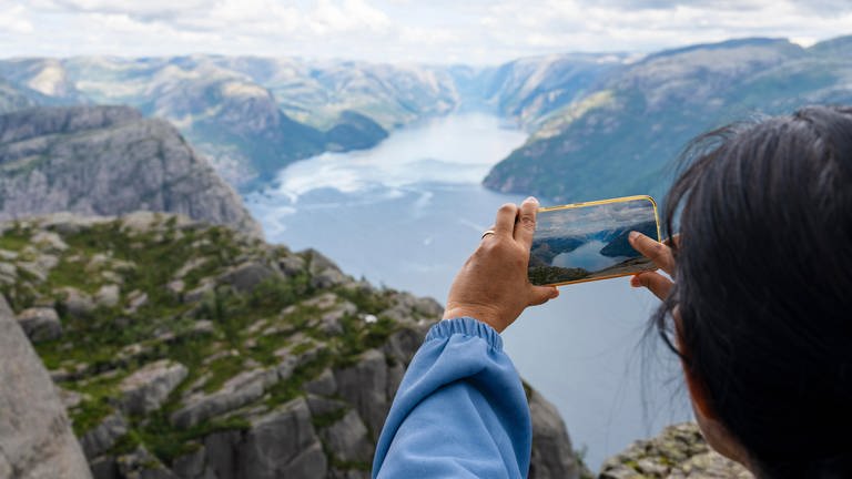 Frau steht auf dem Berg mit dem Felsen Preikestolen mit aussicht auf den Fjord Lysefjord und macht ein Foto mit dem Smartphone. (Foto: picture-alliance / Reportdienste, picture alliance / CHROMORANGE | Michael Bihlmayer)