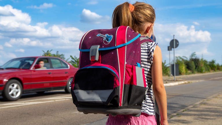 Ein Mädchen mit Ranzen läuft zur Schule, ein Auto fährt vorbei. Kinder sollten auf ihrem Schulweg möglichst nicht allein laufen.