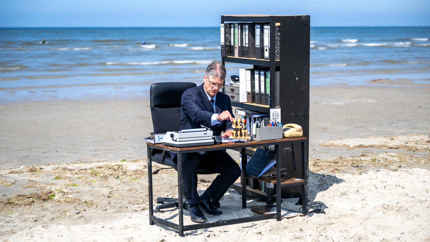 Workation – Ein Mann sitzt am Strand am Schreibtisch (Foto: picture-alliance / Reportdienste, picture alliance/dpa | Sina Schuldt)