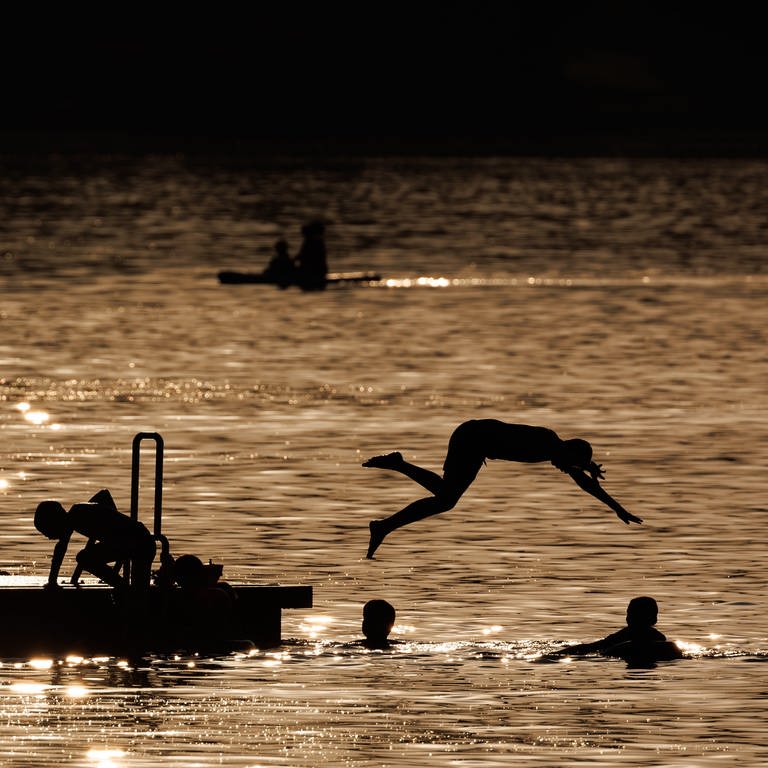 Ausflügler springen am Abend von einem Schwimmsteg in den Igelsbachsee.